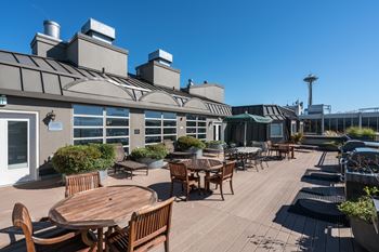 A patio with wooden tables and chairs is surrounded by a building with a grey roof.